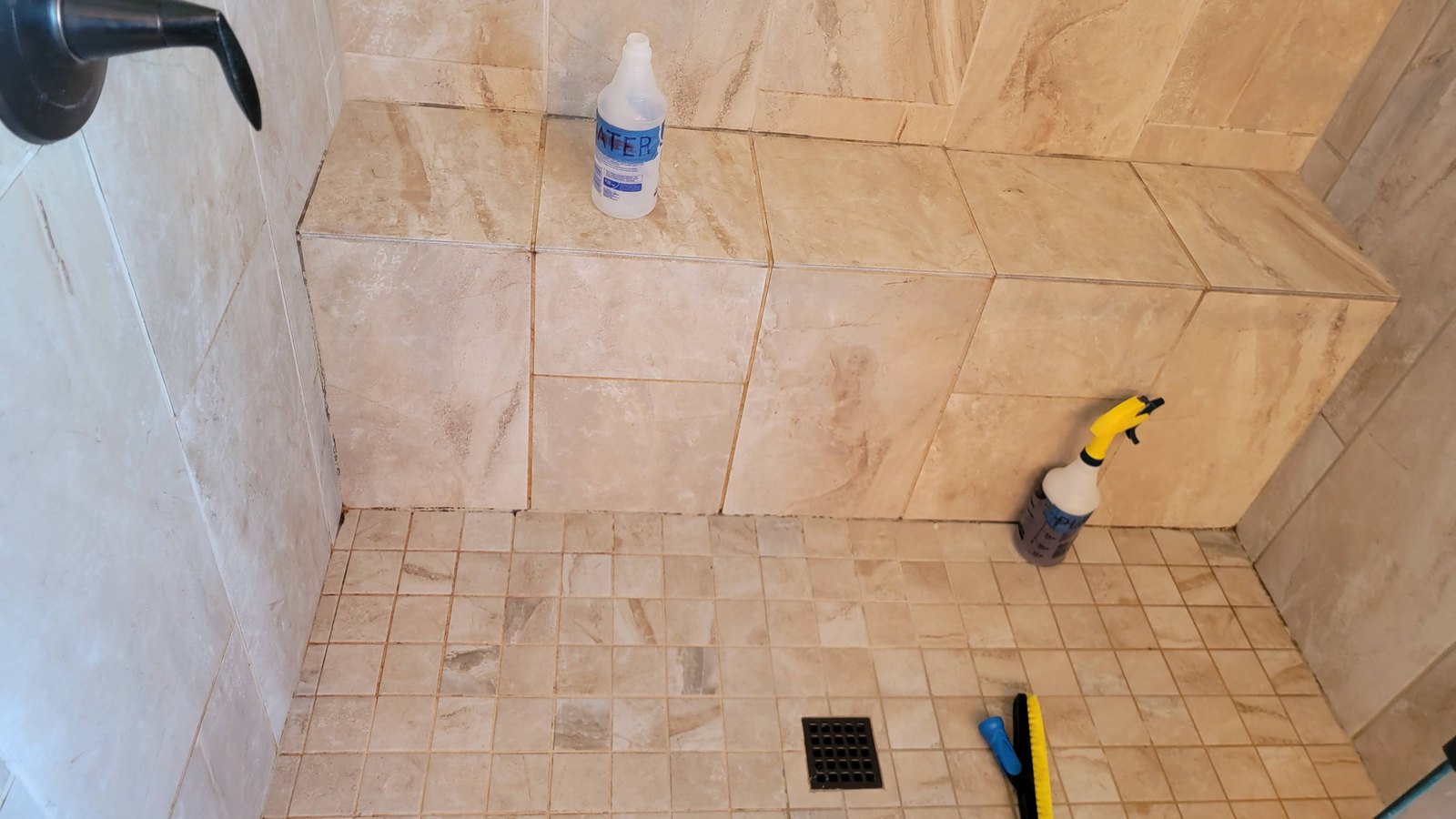 Travertine shower in Kansas City part way through a grout cleaning, mosaic floor and grout lines darkened, brush and sprayer on the floor